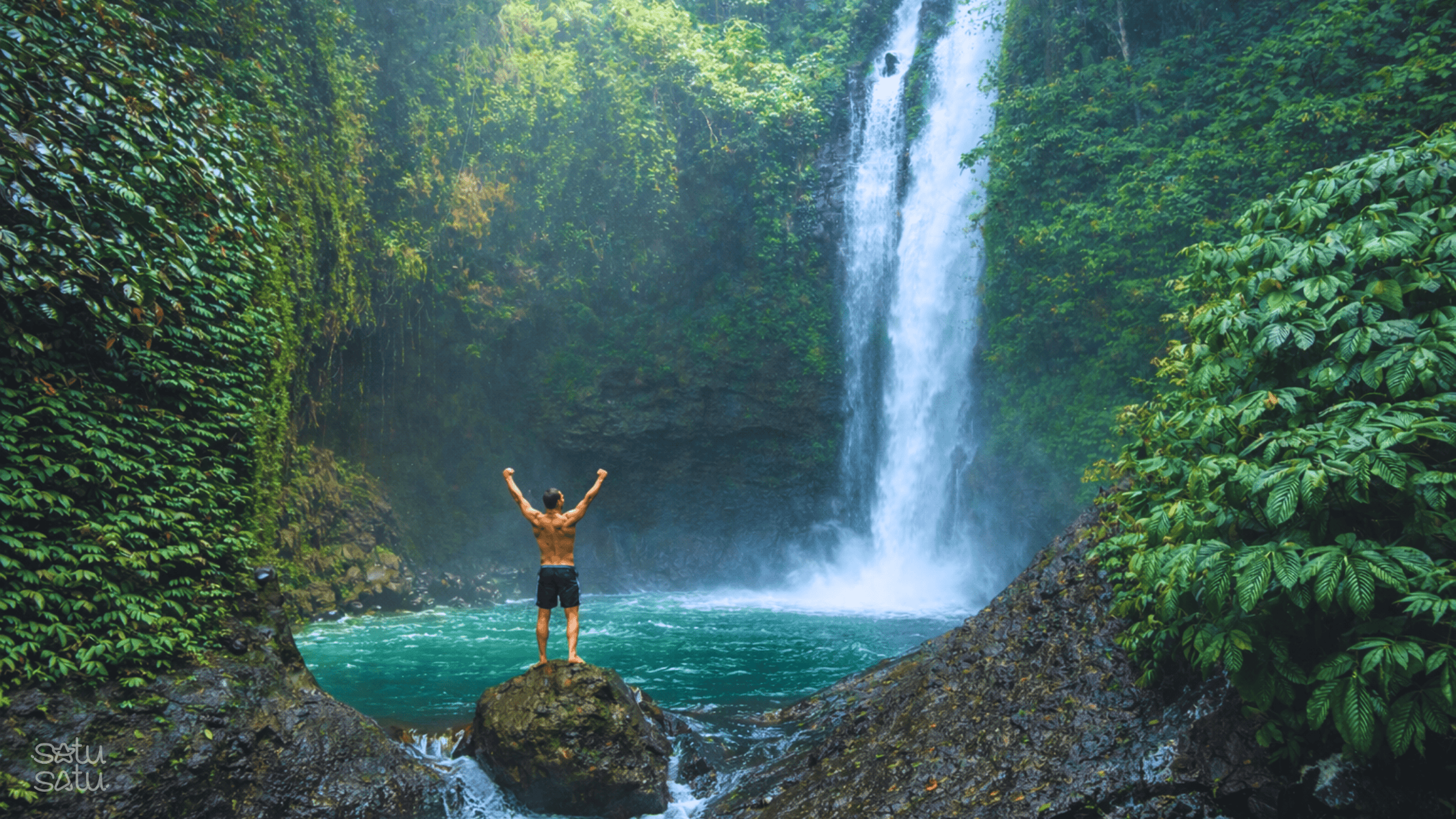 A traveler standing on a rock facing Sambangan Waterfalls surrounded by lush tropical forest in Bali.
