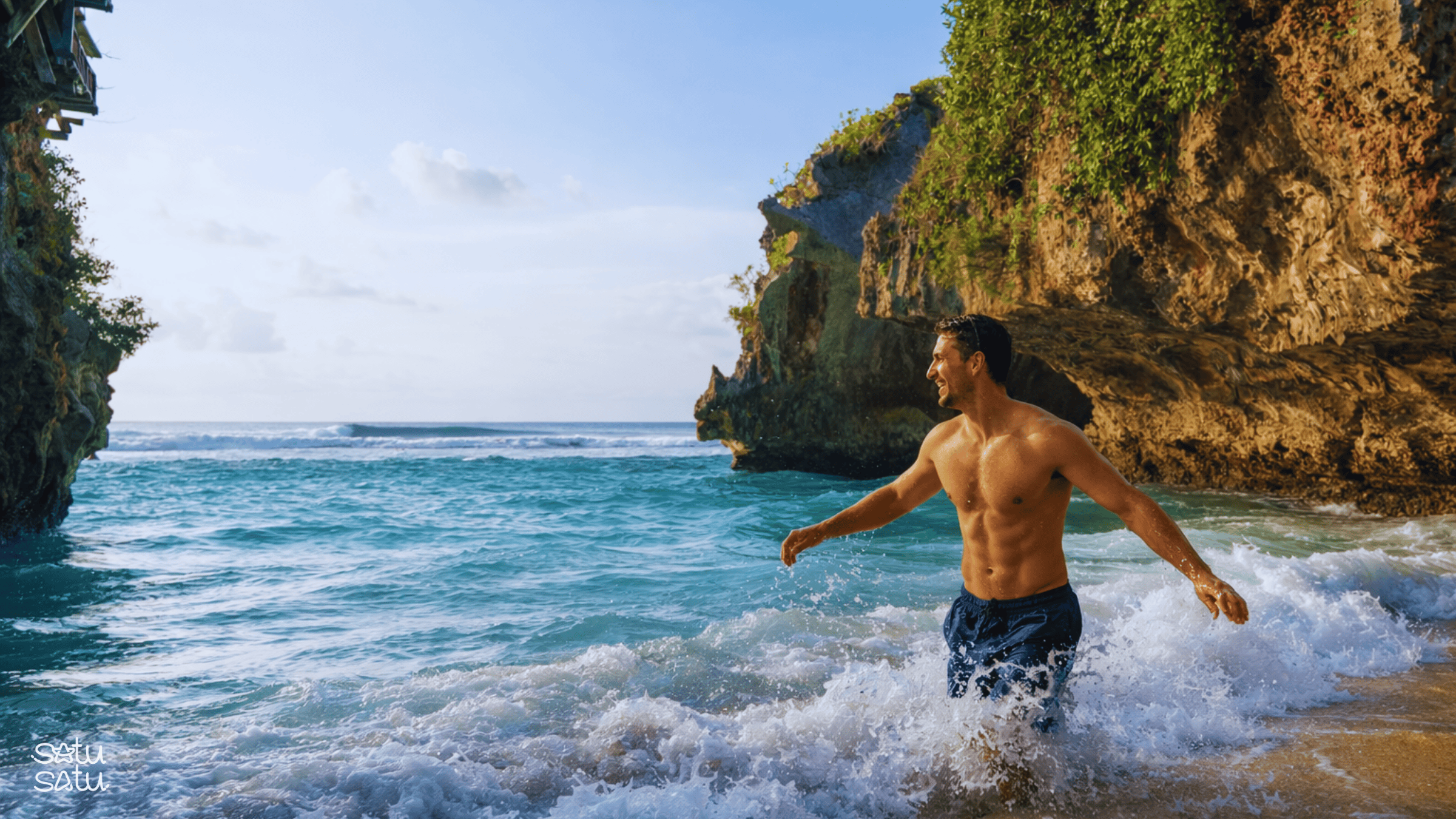 A man walking through turquoise waves at Suluban Beach in Bali, surrounded by dramatic limestone cliffs and a hidden coastal cove.